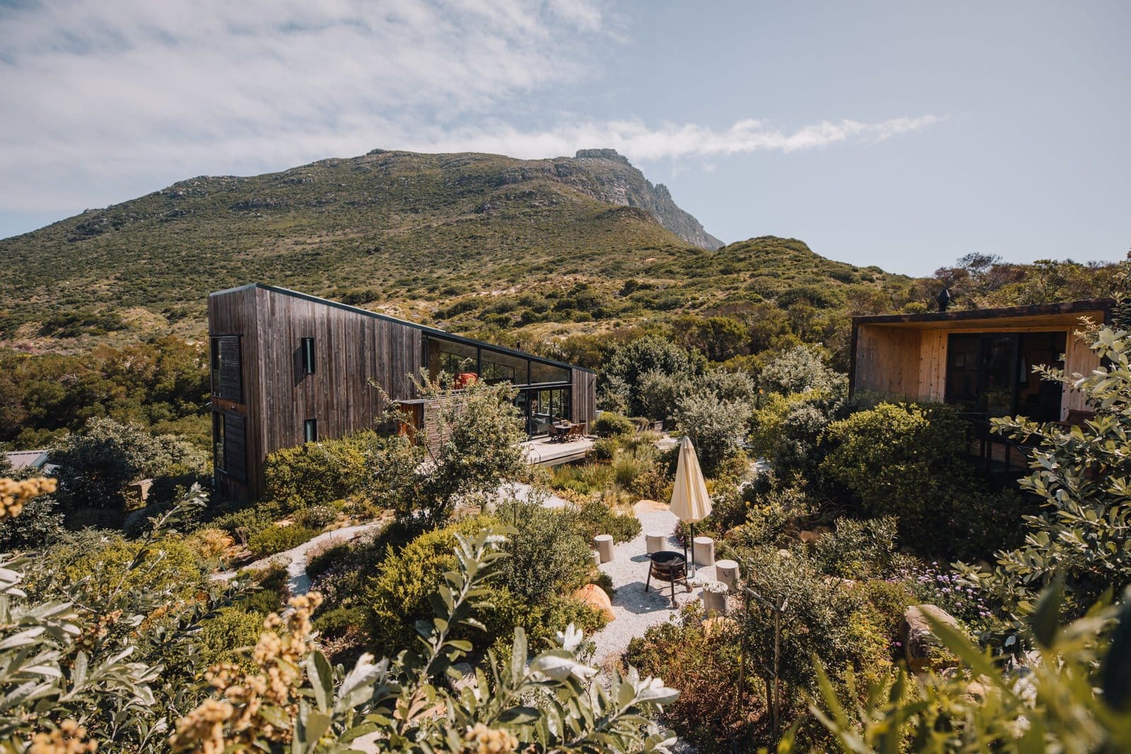 A low timber cabin with an outdoor terrace, nestled in fynbos on a Cape Town mountainside below a mountain ridge.